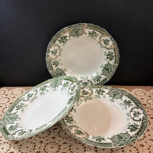 May include: Three white ceramic serving bowls with a green and gold floral and landscape design. The bowls are oval and round, with scalloped edges. The bowls are arranged on a cream-colored lace tablecloth.
