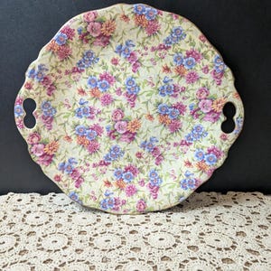 May include: A floral patterned serving tray with a scalloped edge and two handle cutouts. The tray features a cream-colored background with a repeating pattern of pink, blue, and purple flowers. The edge is trimmed with gold.