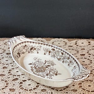 May include: An oval ceramic serving dish with a cream-colored base and brown floral patterns. The interior features a central floral bouquet and a decorative border. The handles on each end have a molded, scalloped, and ribbed design.