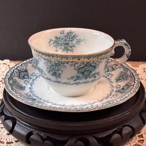 May include: A white porcelain teacup and saucer set featuring a teal floral and scroll pattern. The rim of the cup and the edges of the saucer are accented with a thin, scalloped gold trim. The handle of the cup also features a matching teal pattern.