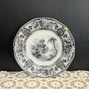 May include: Vintage small plate with a black and white floral and landscape design. The plate features a central scene of a building and palm tree, surrounded by a floral border. The plate has a slightly irregular shape and shows signs of wear.