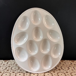 May include: An egg-shaped white ceramic serving platter featuring twelve recessed oval indentations arranged in a structured pattern. The platter has a smooth, glossy finish and a clean, simple design intended for holding and displaying individual eggs.