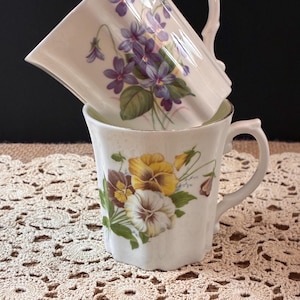 May include: Two white ceramic teacups stacked together. The top cup features a purple violet floral design, while the bottom cup showcases a yellow and white pansy floral pattern. Both cups have curved handles.