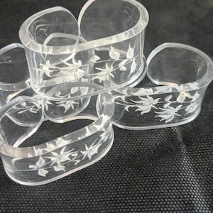 May include: Clear acrylic napkin rings with a floral design. The napkin rings are in a variety of shapes and sizes, and are arranged on a dark surface. The floral design is etched into the acrylic.