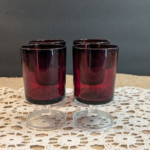 Op de afbeelding: Vier robijnrode wijnglazen. De glazen hebben een heldere steel en voet, met een donkerrode kom. De glazen zijn gerangschikt op een wit kanten tafelkleed. De achtergrond is donkergrijs.