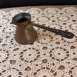 May include: A copper coffee pot with a wide base, a narrow neck, and a flared rim. It features a long, dark, textured metal handle attached to the side with a brass-colored mount. The interior of the pot is dark, and the exterior has a metallic finish.