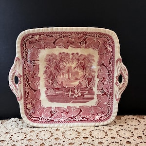May include: A square ceramic serving platter with a cream-colored base and a deep red transferware pattern. The center features a landscape with figures, while the border displays a detailed grape leaf and vine motif with two side handles.