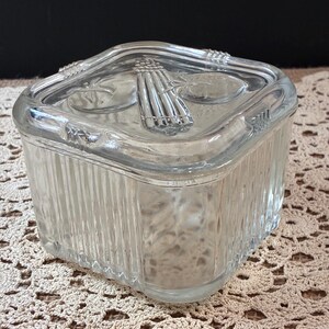 May include: A clear, square glass jar with a lid. The jar has vertical ribbed sides and a decorative design on the lid. The lid features a central design of a bundle of wheat or similar stalks.