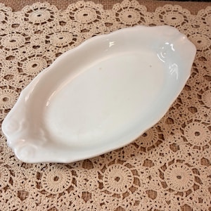 May include: An oval-shaped white ceramic serving dish with a smooth, glossy finish. The edges of the dish feature a subtle, raised scalloped design with decorative embossed floral details at both ends, creating a refined and classic appearance.