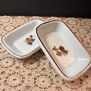 May include: Two rectangular white ceramic dishes with rounded corners, featuring a brown floral motif at the center of each basin and a thin brown painted rim along the top edge. The dishes have a smooth, glossy finish and a shallow depth.