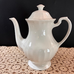 May include: A white ceramic teapot featuring a fluted base, a curved handle, a long spout, and a matching lid with a small knob. The teapot has a smooth, glossy finish and a classic shape, standing upright with a wide body and a narrow neck.