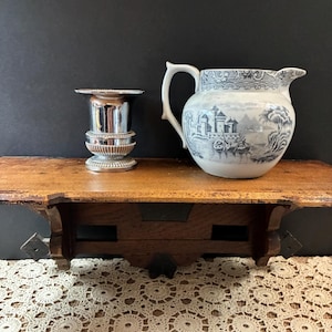 Può includere: Una coppa in metallo argentato con bordo svasato e base arrotondata accanto a una brocca in ceramica bianca con una stampa blu che raffigura un castello, una barca sull'acqua e alberi. Entrambi gli oggetti sono posti su una mensola in legno.