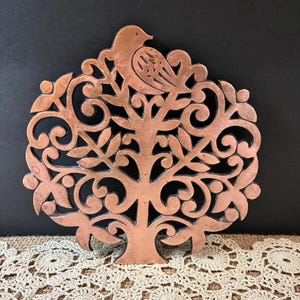 May include: A copper-colored metal wall plaque featuring a cutout design of a tree with swirling branches and leaves. A bird is perched at the very top of the tree. The metal has a warm, metallic finish with intricate scrollwork details.