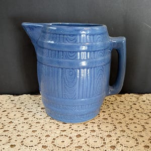 May include: A blue ceramic pitcher with a barrel-like design. The pitcher has a spout and a handle. The surface has a wood grain texture. The pitcher is sitting on a cream-colored lace doily.