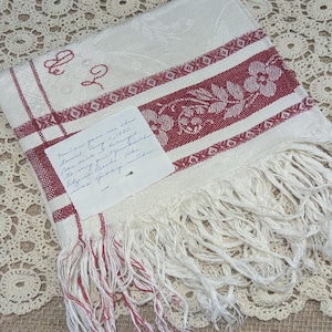 May include: Cream-colored vintage linen towel with red floral and geometric border designs. Features fringed edges and embroidered initials. A handwritten note is attached.