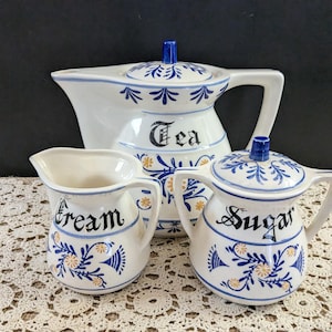 Puede incluir: Juego de té de cerámica blanca con diseños florales azules y amarillos. El juego incluye una tetera con la etiqueta "Tea", una crema con la etiqueta "Cream" y un azucarero con la etiqueta "Sugar".