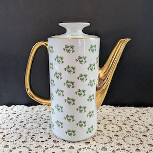 May include: A white porcelain coffee pot featuring a repeating green shamrock pattern across the cylindrical body. The vessel includes a gold-toned handle and a matching gold-toned spout, with a white lid topped by a flared finial and gold trim.
