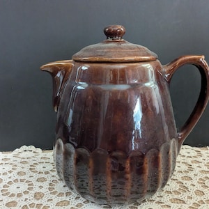May include: A brown ceramic teapot with a lid and handle. The teapot has a glossy finish and a scalloped design around the base. The spout is pointed, and the handle is curved.