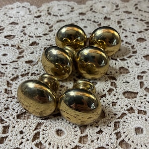 May include: A collection of six shiny, gold-colored cabinet knobs. Each knob has a rounded, spherical shape with a short, cylindrical base. The knobs are arranged on a cream-colored, crocheted doily, creating a vintage aesthetic.