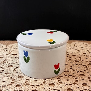 May include: A white ceramic lidded jar with a speckled finish, featuring hand-painted floral motifs in blue, yellow, and red. The cylindrical container has a matching flat lid, both decorated with small, simple flower designs with green stems.