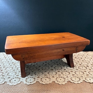 May include: A rectangular wooden stool with a smooth, warm-toned finish. The stool features a flat top surface and four sturdy, slightly angled legs. The wood grain is visible across the top, and the construction includes visible joinery details.