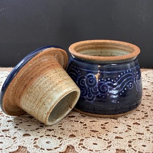 May include: A ceramic butter keeper with a two-piece design. The base is a deep blue with a carved, swirling wave pattern and a tan rim. The lid features a matching blue top and a tan, unglazed cylindrical interior for holding butter.