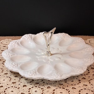 May include: A white ceramic oyster plate with twelve oval indentations arranged in a circular pattern. The plate features embossed grape cluster motifs along the scalloped edge and a central silver-toned metal handle for carrying.