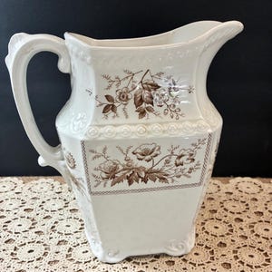 May include: A white ceramic pitcher featuring a brown floral transferware pattern. The vessel has a square-shaped body with rounded corners, a curved handle, and a pouring spout. Embossed decorative bands encircle the upper section of the pitcher.