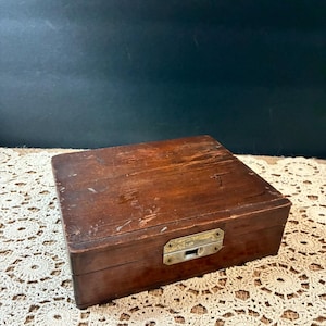 May include: A rectangular wooden box with a dark brown finish and visible surface wear. The box features a centered, weathered metal latch mechanism on the front panel, designed for securing the lid to the base of the container.