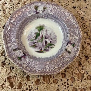 May include: A round ceramic plate featuring a central transferware scene of stone ruins, trees, and figures. The rim is decorated with a repeating purple and white pattern of scrollwork and smaller landscape vignettes in a classic transferware style.