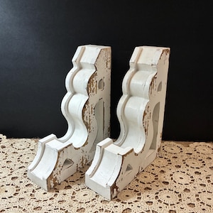 May include: A pair of white wooden corbels featuring a distressed finish that reveals brown wood underneath. Each corbel has a curved, scroll-like profile with decorative cutouts and a flat top surface, standing upright in a matching set.