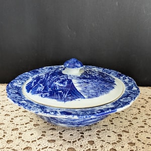 May include: A blue and white ceramic serving dish with a lid. The dish features a landscape design on the lid and rim. The dish is round and has a scalloped edge. The knob on the lid is blue.