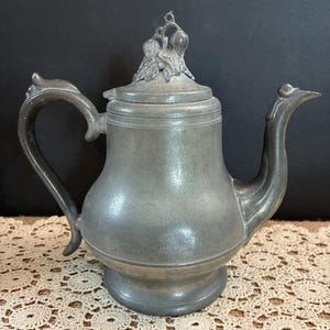 May include: A metallic gray pewter teapot with a curved handle and a spout. The lid features a decorative finial shaped like a cluster of fruit and leaves. The teapot has a rounded body, a wide base, and a smooth, matte finish throughout the surface.
