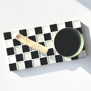 May include: A rectangular tray with a black and white chequered design. A dark green cup and a piece of light-coloured wood are on the tray. The tray is made of small square tiles.