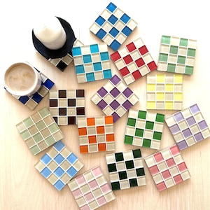 Könnte beinhalten: Eine Sammlung farbenfroher quadratischer Mosaik-Untersetzer in verschiedenen Farben, darunter Blau, Rot, Grün und Lila. Einige Untersetzer haben ein Schachbrettmuster. Eine Tasse Kaffee und eine Kerze sind ebenfalls vorhanden.
