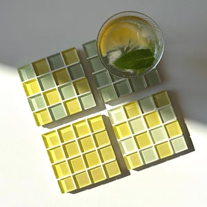 Puede incluir: Cuatro posavasos cuadrados con un diseño de mosaico en tonos amarillos y verdes. Un vaso de agua con hielo, una rodaja de limón y una hoja de menta se encuentra en uno de los posavasos. Los posavasos están dispuestos sobre una superficie blanca.