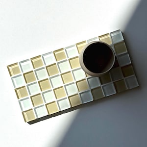 May include: A small, beige ceramic cup sits on a rectangular mosaic tile tray. The tray is made of small, square tiles in shades of beige, white, and light green. The cup contains a dark liquid.