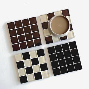 Puede incluir: Cuatro posavasos cuadrados de azulejos en tonos marrón, negro y crema. Un posavasos sostiene una taza marrón claro. Los posavasos están dispuestos sobre una superficie blanca. Los posavasos están hechos de pequeños azulejos cuadrados.