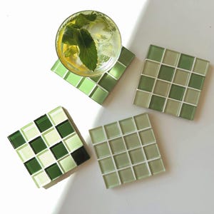 May include: Four square glass tile coasters in shades of green. One coaster holds a glass of a refreshing drink with mint and ice. The coasters are arranged on a white surface, with a bright light shining on them.