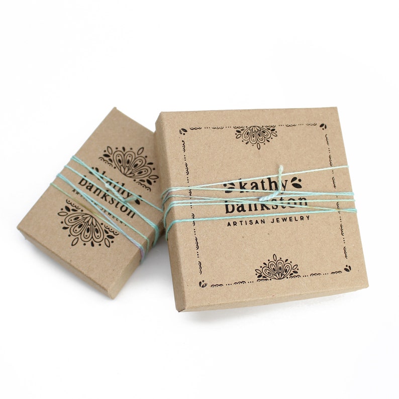 May include: Two brown cardboard boxes with a decorative design and the text "kathy bankston artisan jewelry" printed on the front. The boxes are tied with a light blue string.