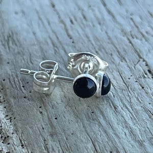 May include: A pair of sterling silver stud earrings featuring round, polished black onyx stones. Each earring has a secure post back with the 925 hallmark clearly visible on the metal backing, showcasing a simple and classic design.