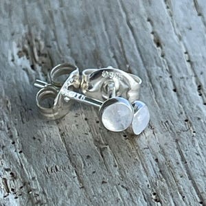 May include: A pair of small, round, silver stud earrings with a light-colored, possibly moonstone, center. The earrings are lying on a weathered, gray wooden surface. The earring posts and backs are visible.