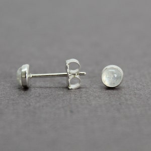 May include: A pair of silver stud earrings with a round, white moonstone gemstone in each earring.