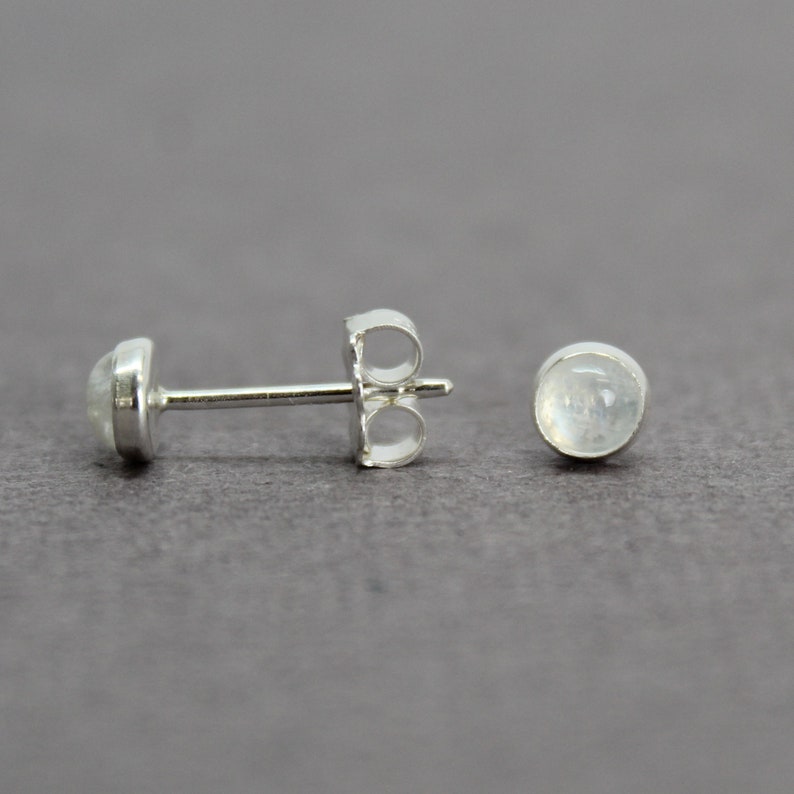 May include: A pair of silver stud earrings with a round, white moonstone gemstone in each earring.