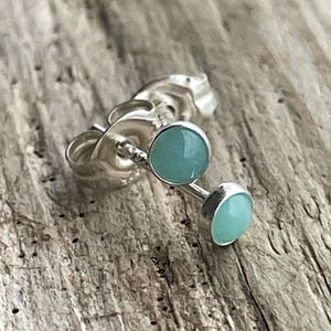 May include: A pair of silver stud earrings featuring small, round, smooth, light blue-green cabochon stones. Each earring has a polished silver post and a matching silver butterfly clutch back, showcasing a simple and clean design.