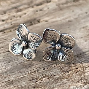 May include: A pair of silver-toned flower stud earrings. Each earring features a detailed, three-dimensional flower design with a small, round center. The earrings are set against a wooden surface.