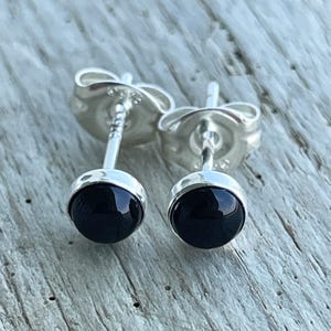 May include: A pair of silver stud earrings with round, polished black stones. The earrings have a simple, classic design, with the black stones set in a bezel setting. The posts and backs are silver. The earrings are displayed on a light-colored wooden surface.