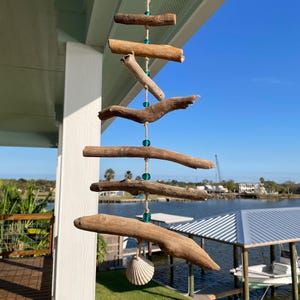 Puede incluir: Móvil artesanal de madera flotante con piezas de madera natural suspendidas por cuerdas. Cuentas turquesas acentúan el diseño. Una concha blanca cuelga en la parte inferior. El móvil se exhibe al aire libre con vista al agua.