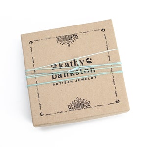Peut inclure: Une bo&icirc;te en carton marron avec un motif d&eacute;coratif en haut et en bas. La bo&icirc;te est attach&eacute;e avec une ficelle bleue et blanche et porte l'inscription "kathy bankston artisan jewelry" sur le devant.