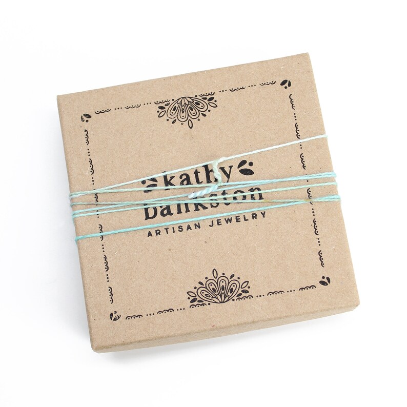 Peut inclure: Une bo&icirc;te en carton marron avec un motif d&eacute;coratif en haut et en bas. La bo&icirc;te est attach&eacute;e avec une ficelle bleue et blanche et porte l'inscription "kathy bankston artisan jewelry" sur le devant.